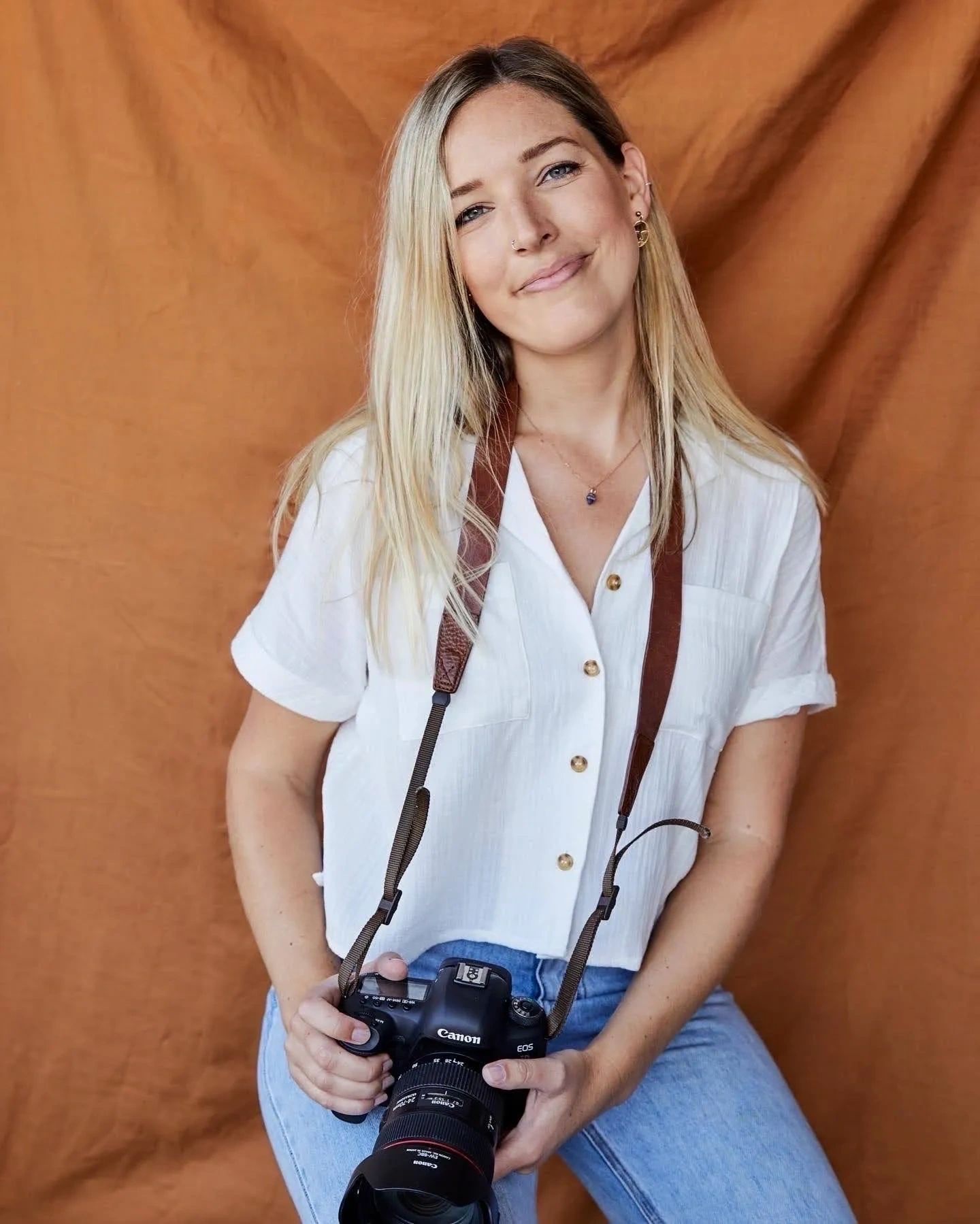 Monica Pronk seated in front of a rust-coloured cloth backdrop, camera in hand.