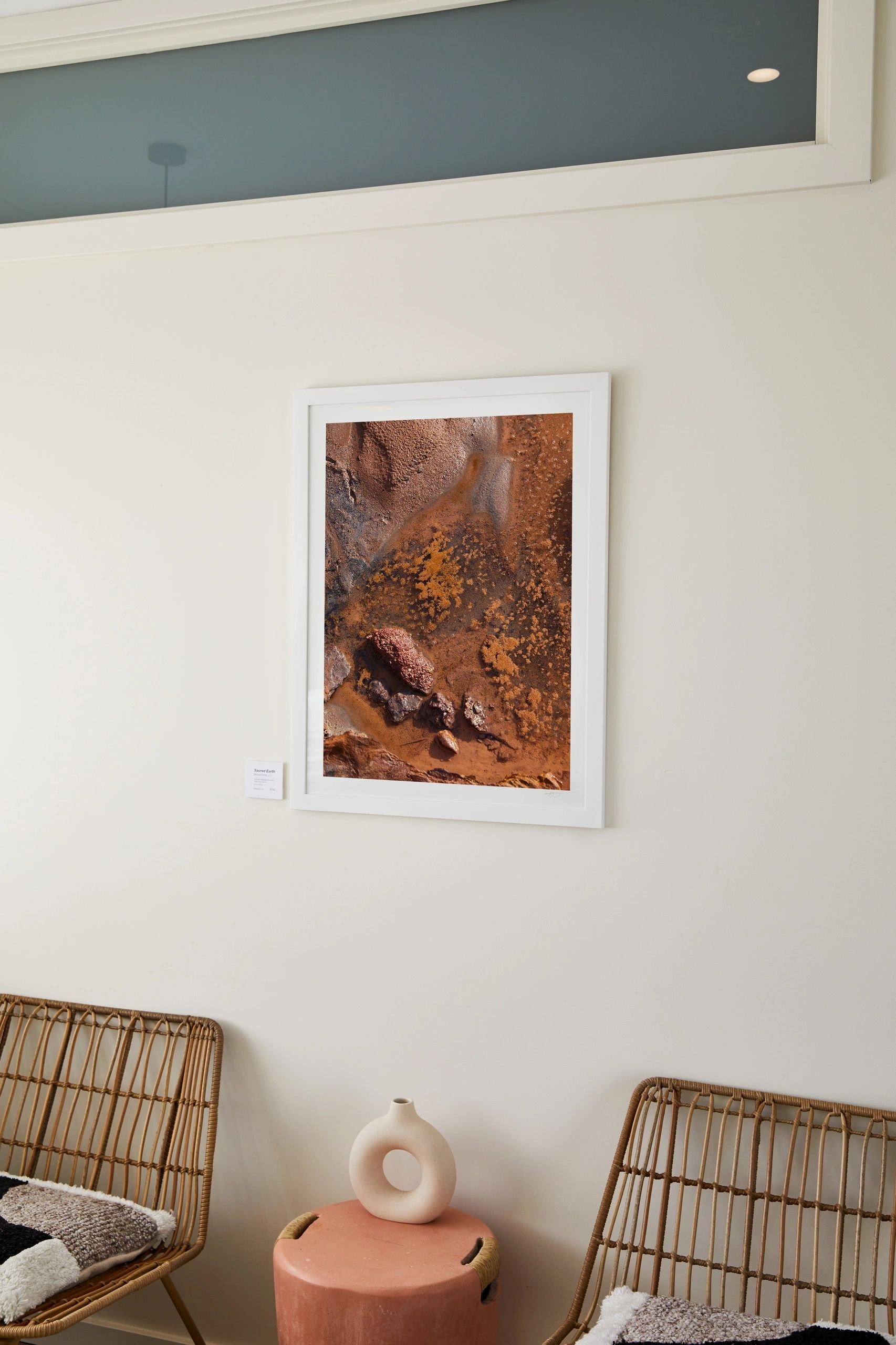 Sacred Earth, a framed print of ochre rock and earth textures, hanging in the Mesa studio above two rattan chairs.